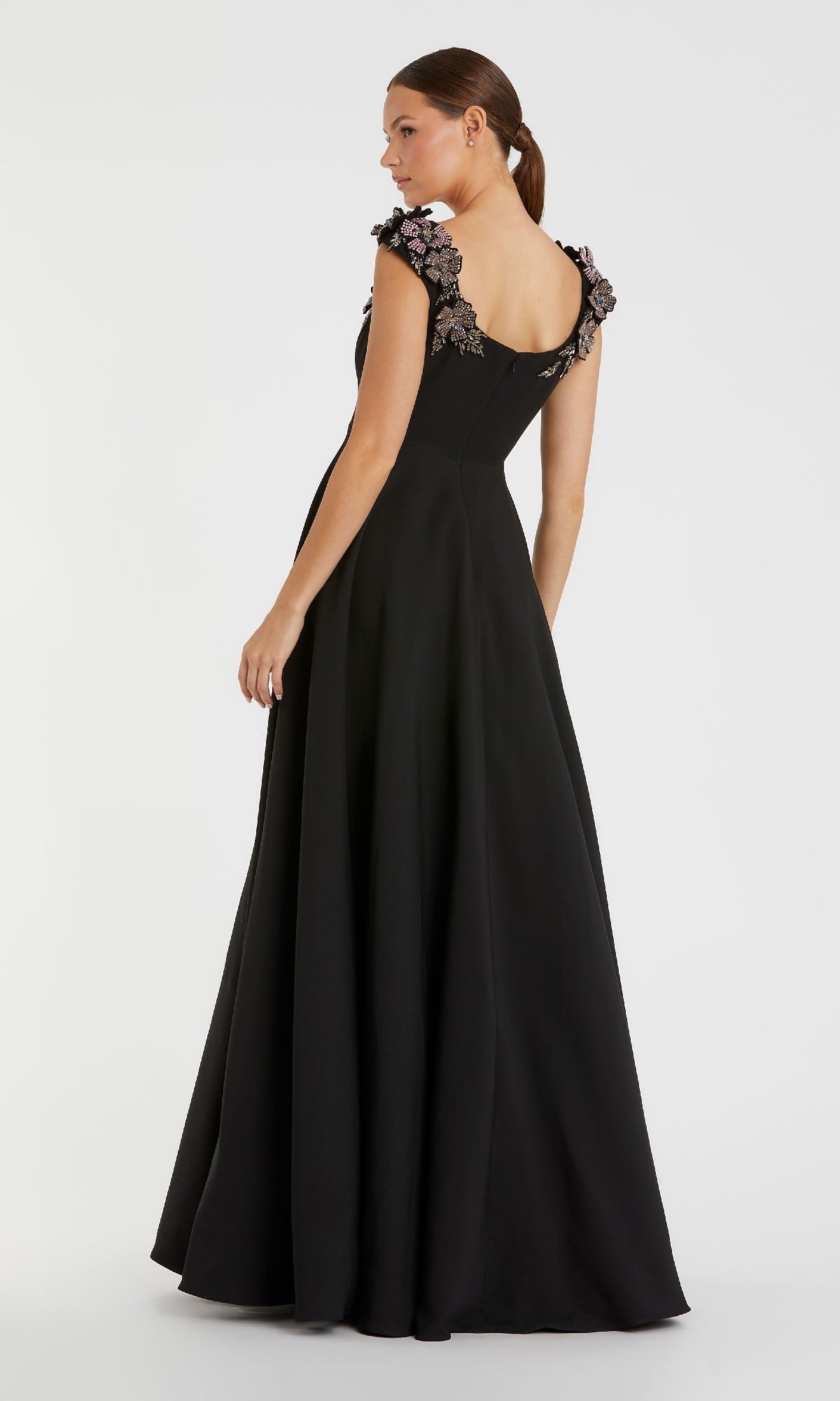 A woman wearing the Mac Duggal 11949 long black A-line formal dress with floral embroidered shoulders stands with her back to a plain white background.